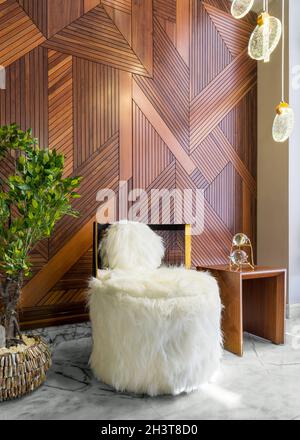 Modern white feather armless chair, small wooden modern table, and planter with green bushes, in a hall with decorated wood cladding wall, and white marble floor Stock Photo