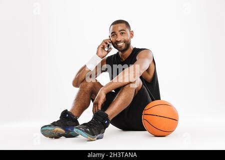 Happy african young basketball player sitting talking on mobile phone isolated on a white background Stock Photo