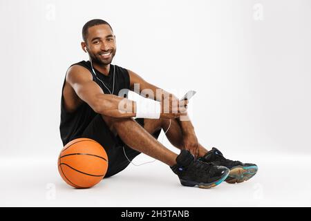 Happy african young basketball player sitting listening to music with earphones and mobile phone isolated on a white background Stock Photo