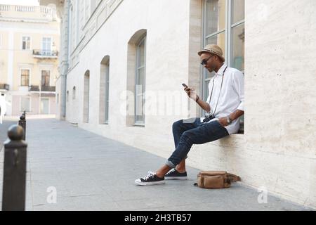Black man sitting on windowsill and using mobile phone on street in european city. Young guy with digital camera wear straw hat, white shirt, jeans, g Stock Photo