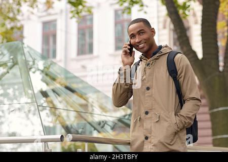 Smiling black male student standing with hand in pocket and talking on mobile phone. Young mulatto man wear parka coat and backpack. Blurred city back Stock Photo