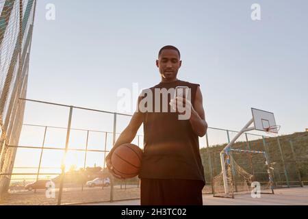Young concentrated african sportsman holding basketball ball and using mobile phone on sports court. Black man wearing sportswear. Urban basketball pl Stock Photo
