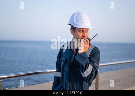 Positive young seaman in hardhat and protective uniform communicating via walkie talkie while standing near railing against sea in port Stock Photo