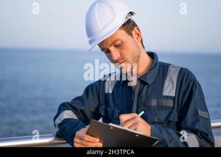 Serious young male port employee in protective uniform and hardhat with clipboard taking notes in documents while standing against sea Stock Photo
