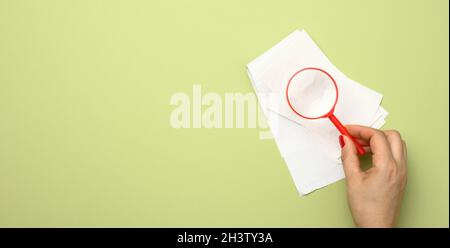 A stack of paper sales receipts and a woman's hand with a red plastic magnifying glass on a green background. The concept of con Stock Photo