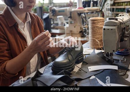 Close-up of shoemaker sitting at her workplace and making new shoes in the workshop, she sewing leather details Stock Photo