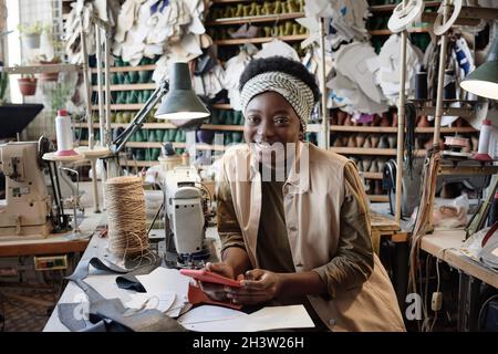Portrait of African female tailor smiling at camera while using mobile phone at her workpalce in workshop Stock Photo