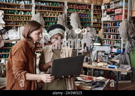 Two dressmakers working online on laptop and discussing the new way of sewing together while working in the workshop Stock Photo