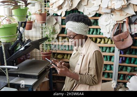 African dressmaker talking online with her customers using mobile phone while standing in the workshop Stock Photo