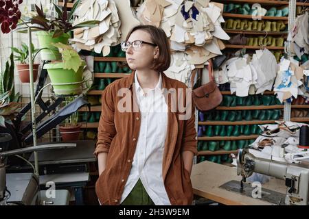 Female shoe designer looking away with pensive sight while standing in the workshop with products in the background Stock Photo