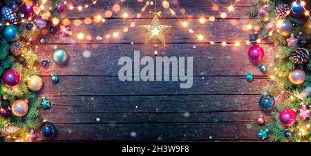 Christmas Decoration - Fir Branches And Baubles Illuminated With String Lights On Dark Wooden Table With Abstract Snowfall Stock Photo