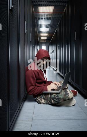 The hacker in a hood sits with the computer among servers, the burglar, face covered with a hood. Storage of data, cloudy services, system breaking Stock Photo