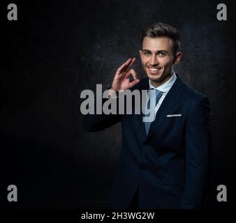 A young male steward, in a suit, smiles, makes a hand gesture everything is ok. Against a dark background. Stock Photo