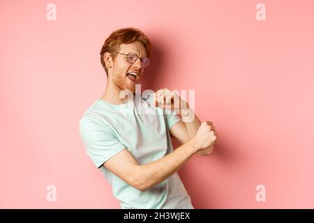 Summer vacation concept. Happy young man with red hair dancing and having fun, singing along music, standing over pink backgroun Stock Photo