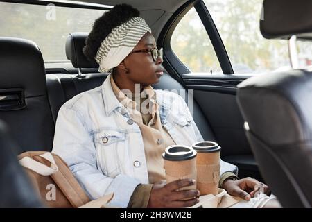 African woman with coffee drinks looking through the window while sitting on back seat of the taxi Stock Photo