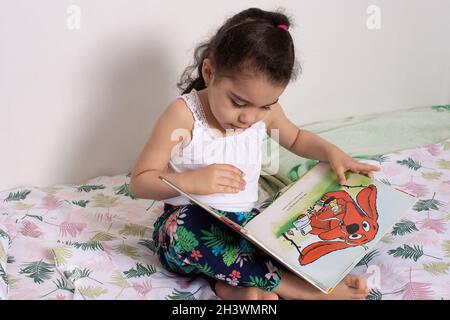 Preschool child at home, 4 year old girl sitting on bed looking at picture book Stock Photo