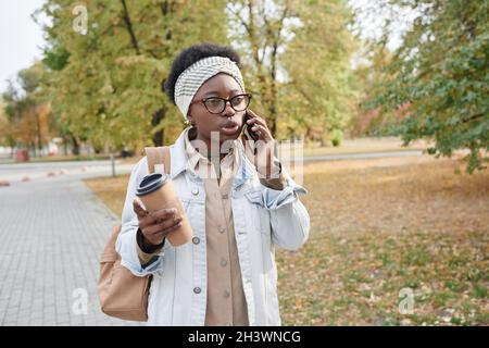 African young woman walking in the park during her coffee break and talking on mobile phone Stock Photo