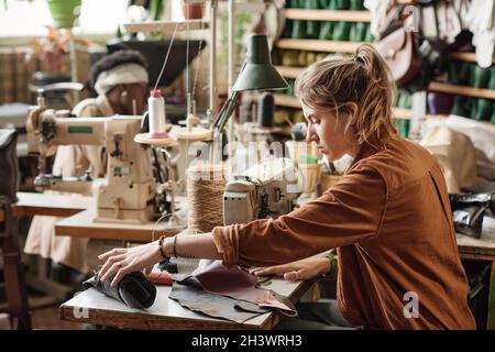Young dressmaker sewing pieces of leather together using sewing machine while working in the factory Stock Photo