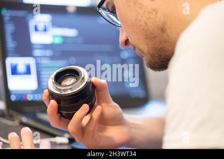 Male photographer has a lens in his hand and is looking to it. Laptop in the blurry background. Stock Photo