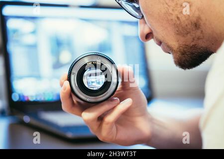 Male photographer has a lens in his hand and is looking to it. Laptop in the blurry background. Stock Photo