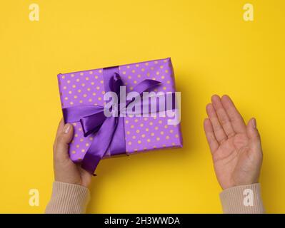 Two female hands are holding a purple gift box on a yellow background, happy birthday concept Stock Photo