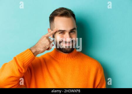 Image of young man in orange sweater, smiling and pointing at head, having an idea, praising good thought, standing over light b Stock Photo