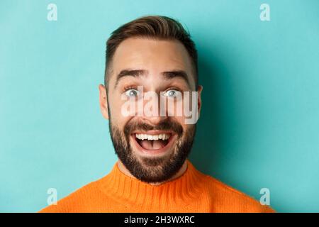 Headshot of handsome caucasian man with beard smiling happy at camera, staring amazed, standing in orange sweater against turquo Stock Photo