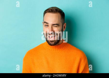 Handsome confident man winking at camera, smiling sassy, standing in orange sweater against blue background Stock Photo