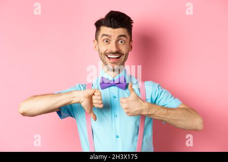 Funny young man smiling and showing thumbs up down, judging something and being indecisive, making choice, standing on pink back Stock Photo