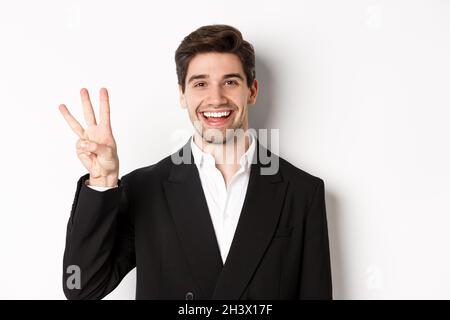 Close-up of handsome businessman in black suit, smiling amazed, showing number three, standing over white background Stock Photo
