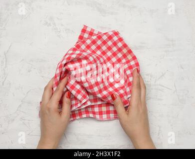 Two female hands hold a crumpled white-red checkered textile kitchen napkin on a white table Stock Photo