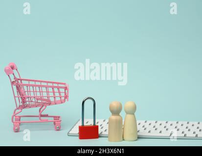 A red metal padlock stands on the keyboard and behind a miniature shopping cart, blue background Stock Photo