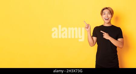 Portrait of cheerful asian man pointing fingers upper left corner, smiling and showing banner satisfied, yellow background Stock Photo