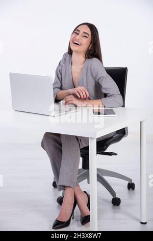 Young caucasian woman with long black hair. She is wearing a grey jumpsuit and sitting at her office.She is in front of her laptop and laughing. Stock Photo