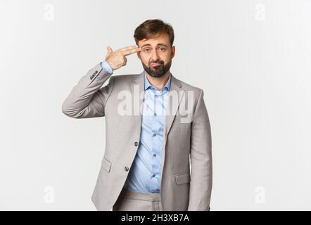 Image of miserable businessman with beard, wearing grey suit, making finger gun sign over head and shooting himself, standing di Stock Photo