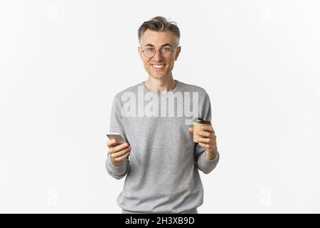 Handsome middle-aged man in grey sweater and glasses, smiling happy, drinking coffee and using mobile phone, standing over white Stock Photo