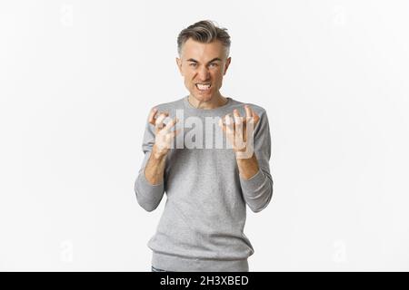 Image of angry middle-aged man cursing someone, looking mad and clenching fists with hatred, standing over white background Stock Photo