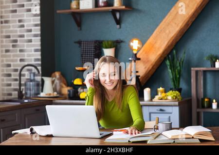 Young smiling woman freelancer sitting at table at home with laptop, making notes Stock Photo