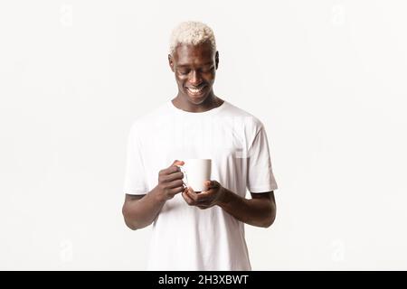 Portrait of relaxed happy african-american blond guy, looking dreamy and smiling at cup of coffee, standing white background Stock Photo