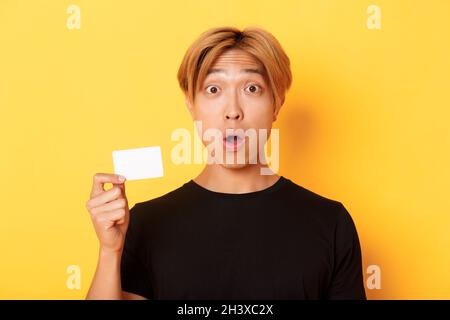 Close-up of surprised gasping asian guy with fair hair, looking amazed and startled as showing credit card, standing yellow back Stock Photo