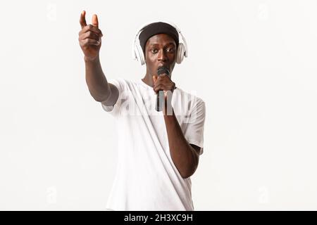 Portrait of sassy and cool african-american guy in wireless headphones, singing into microphone, standing white background Stock Photo