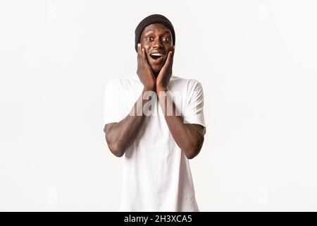 Portrait of happy surprised young african-american guy, looking amazed and cheerful, gasping fascinated, holding hands on cheeks Stock Photo