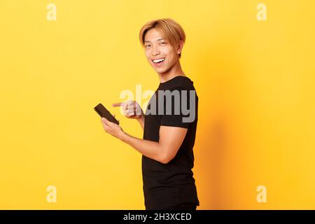 Portrait of pleased smiling handsome asian man, standing in profile and pointing finger at smartphone, recommend app, standing y Stock Photo
