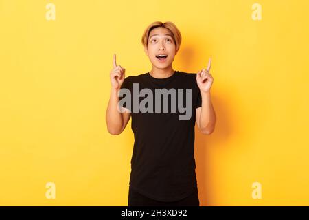 Impressed good-looking asian blond guy, wearing black t-shirt, looking and pointing fingers up excited, yellow background Stock Photo