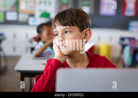 Sad caucasian boy with laptop sitting on his desk in the class at elementary school Stock Photo