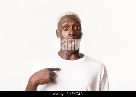 Portrait of startled, african-american blond guy looking surprised as pointing at himself with dropped jaw, standing over white Stock Photo