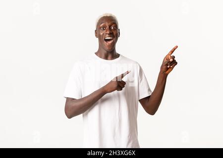 Portrait of surprised and excited african-american guy, looking fascinated and pointing fingers upper right corner, white backgr Stock Photo