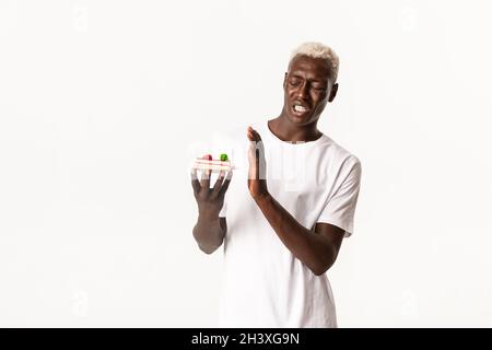 Portrait of disgusted handsome african american young blond man, unwilling eat dessert, cringe from disgusting cake, standing wh Stock Photo