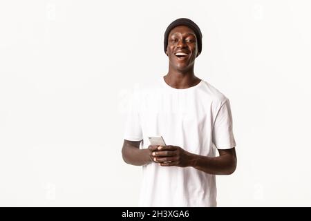 Portrait of carefree attractive african-american guy in beanie laughing and smiling, using mobile phone application, white backg Stock Photo