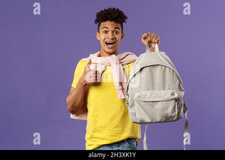 Portrait of cheerful young handsome male student, man recommending backpack, holding rucksack and show thumbs-up, bought new equ Stock Photo